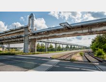 Industrieanlage mit erhöhten Rohrleitungen aus Metall, die über eine Straße führen, neben denen sich Bahngleise und vereinzelte Bäume befinden. Im Hintergrund sind Zäune, Gebäude und ein bewölkter Himmel zu sehen.