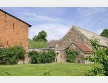 Hof mit historischen Gebäuden aus Naturstein und rotem Backstein, umgeben von niedrigen Mauern und bewachsenen Fassaden. Im Vordergrund eine große Rasenfläche, im Hintergrund Bäume und ein klarer Himmel.