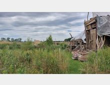 Holzverschlag mit einer improvisierten Konstruktion aus Ästen und Planen, umgeben von hohem Gras und wildem Bewuchs; im Hintergrund eine flache, ländliche Wiesenlandschaft unter bewölktem Himmel.