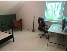 Ein einfach eingerichtetes Zimmer mit weißen Holzböden und schräger Decke. Links ein Metallbett und ein brauner Lounge-Sessel, rechts ein Schreibtisch mit Bürostuhl und Monitor, daneben ein Fenster mit Blick auf grüne Bäume.