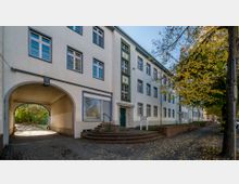 Mehrstöckiges Wohngebäude mit weißer Fassade und blauen Fensterrahmen; ein Torbogen führt zu einem Innenhof. Rechts im Bild steht ein Baum mit gelben Herbstblättern, und der Gehweg ist mit Laub bedeckt.