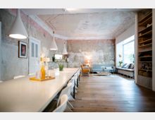 Heller Raum mit einem langen Esstisch und modernen, weißen Hängelampen, ergänzt durch eine Lounge-Ecke mit einem Sofa und Sesseln. Die Wände sind teilweise unbehandelt mit sichtbarem Putz, während Holzfußboden und große Fenster für ein warmes und helles Ambiente sorgen.