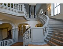 Historisches Treppenhaus mit geschwungenem Geländer, verzierten Balustern und Marmorsäulen. Der Raum ist geprägt von hell gestrichenen Wänden, Rundbögen, großen Fenstern und eleganter Beleuchtung.