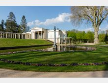 Weißes klassizistisches Gebäude mit Säulen und Satteldach in einer Parkanlage. Im Vordergrund ein runder Teich, umgeben von Blumenbeeten, grünem Rasen und einem großen Baum.