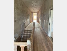 Ein unfertiger Flur in einem Gebäude mit abgenutzten Wänden und einem Holzboden. In der Mitte führt eine Holztreppe nach unten, und am Ende des Flurs befindet sich eine Tür mit einer verglasten Oberseite, durch die Tageslicht einfällt.