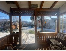 Blick von einer überdachten Holzveranda mit verzierten Geländern auf einen schneebedeckten Garten. Im Hintergrund sind Bäume, ein Gartenhaus sowie mehrere Wohngebäude zu sehen, während im Vordergrund Gartenmöbel und Dekorationen die Veranda schmücken.