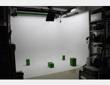 Professionelles Fotostudio mit einem weißen Hintergrund, mehreren grünen Boxen auf dem Boden und Beleuchtungseinrichtungen an der Decke. An den Seiten befinden sich Regale mit Equipment und Zubehör.