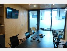 Konferenzraum mit Holzwänden, einem großen schwarzen Tisch und schwarzen Stühlen. Im Raum befinden sich ein Flachbildschirm an der Wand, Glaswände mit teilweise undurchsichtiger Folierung und Blick auf einen angrenzenden Arbeitsbereich.