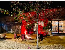 Ein gepflasterter Innenhof bei Nacht mit einem beleuchteten Baum im Vordergrund und einer rot angestrahlten Backsteinfassade im Hintergrund. Vor der Fassade stehen Gartenstühle um eine Feuerstelle, daneben ein hölzerner Tisch und ein alter Handkarren; durch ein verglastes Tor sind ein beleuchteter Innenraum und Dekorationen sichtbar.