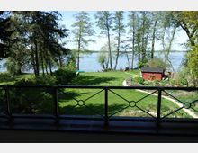 Ein Blick von einem Balkon auf einen gepflegten Garten mit grünem Rasen, Bäumen und einem kleinen roten Gartenhaus. Im Hintergrund ist ein See zu sehen, umgeben von weiteren Bäumen.