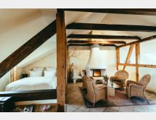 Dachzimmer mit sichtbaren Holzbalken, einer Schlafnische mit Doppelbett links und einer Sitzecke mit geflochtenen Sesseln um einen Glastisch. Im Hintergrund ein weißer Kamin und eine Wand mit Baumdekor.