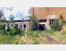 Verlassene Fabrikgebäude mit verwitterten Ziegelmauern, zerbrochenen Fenstern und Graffiti, umgeben von wuchernder Vegetation und hohem Gras; im Vordergrund ein schmaler Trampelpfad.