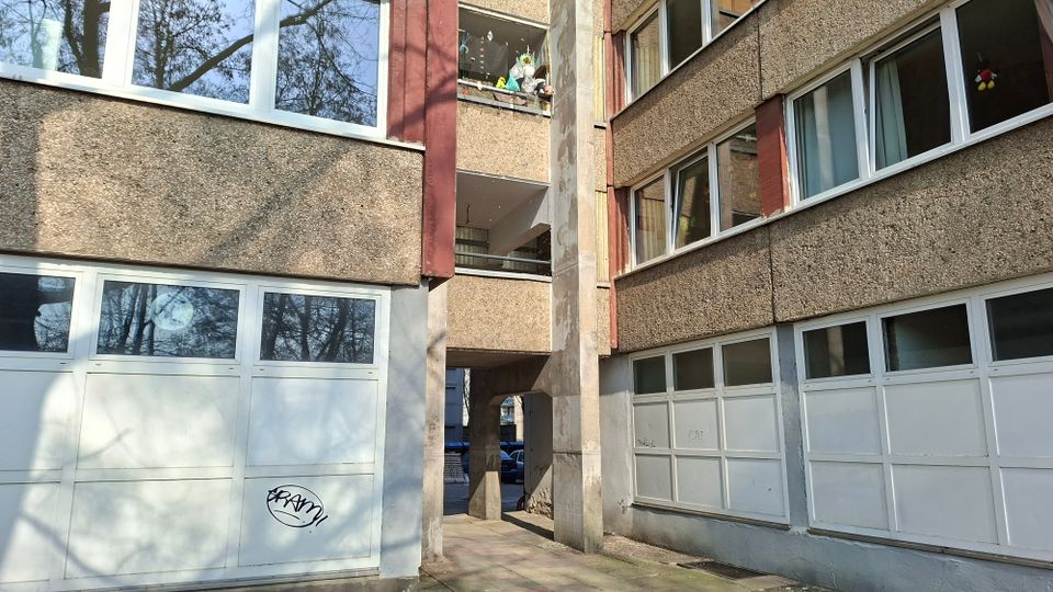 Innenhof zwischen zwei mehrstöckigen Wohnblocks mit rauer Betonfassade, weißen Fensterbändern und roten vertikalen Fassadenelementen. In der Mitte führt ein überdachter Durchgang unter dem Gebäude hindurch; unten sind große weiße Garagentore und seitlich Balkone sichtbar.
