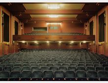 Innenraum eines Theatersaals mit mehreren Reihen gepolsterter Sitze, einer Empore im Hintergrund und warm beleuchteten Deckenlampen. Die Gestaltung umfasst Holzverkleidungen und seitliche Vorhänge, die den Raum flankieren.