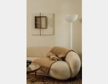 Modernes Wohnzimmer mit einer beigen, geschwungenen Couch, die mit einer braunen Decke dekoriert ist. Im Hintergrund eine minimalistische Wandkunst aus Holz und Metall sowie eine weiße Stehleuchte; ein runder Glastisch steht im Vordergrund.