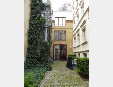 Hof eines Wohngebäudes mit Kopfsteinpflaster, einer efeubewachsenen Wand und einer metallenen Außentreppe links. Im Hintergrund befindet sich eine gelbe Backsteinfassade mit großen Fenstern und einer roten Tür, umgeben von weiteren Gebäuden.