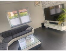 Modernes Wohnzimmer mit großem Ecksofa vor einem Doppelfenster mit Plissees sowie einem Glas-Couchtisch auf dunklen Fliesen. Gegenüber befindet sich eine TV-Wand mit grauer Steinoptik, wandmontiertem Fernseher und weißem Lowboard, daneben eine Zimmerpflanze.