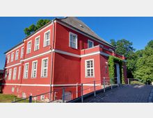 Rotes zweistöckiges Gebäude mit weißen Fensterrahmen und Gesimsen, bedeckt von einem grauen Ziegeldach. Die Umgebung ist grün, mit Bäumen im Hintergrund, und ein gepflasteter Weg führt zum Eingang, der von Kletterpflanzen umrahmt ist.