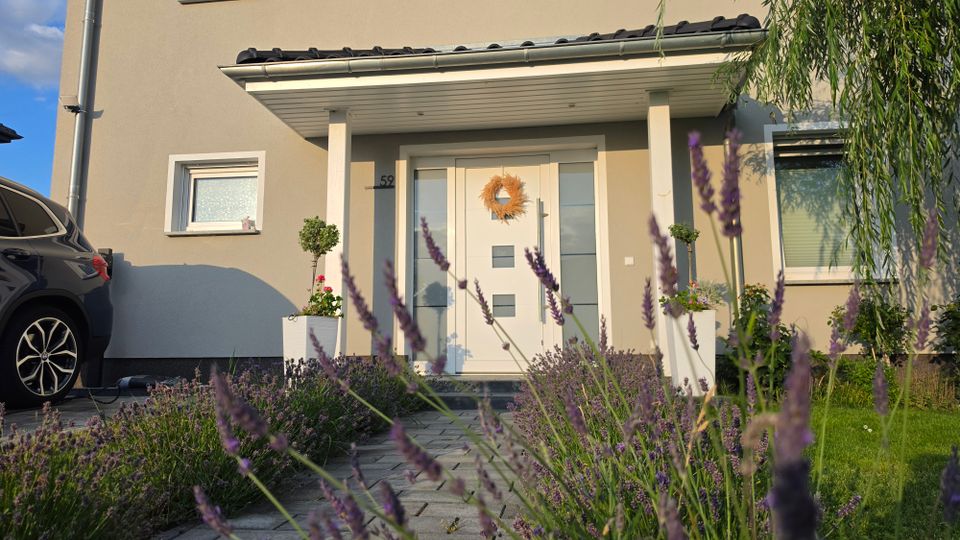 Eingangsbereich eines modernen Wohnhauses mit überdachtem Vorbau, weißer Haustür mit Glaseinsätzen und zwei Säulen. Davor führen Pflastersteine durch einen Vorgarten mit Lavendel und Pflanzkübeln; links ist ein geparktes Auto am Rand der Einfahrt zu sehen.