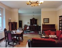 Wohnraum im klassischen Stil mit Holzmöbeln; im Vordergrund ein Esstisch mit Stühlen, rechts ein rotes Sofa und ein Klavier. Im Hintergrund befinden sich eine Anrichte und weitere Möbelstücke, darunter Vitrinen und ein Teppich.