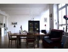 Wohnraum mit hellen Wänden und Holzboden, zentral ein Esstisch aus Holz mit verschiedenen Stühlen. Im Hintergrund ein dunkler Glasschrank, daneben eine Stehlampe und kleinere dekorative Elemente. Auf der rechten Seite mehrere große Fenster, die Tageslicht hereinlassen.