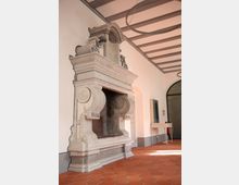 Klassische Innenaufnahme eines Raumes mit auffälligem Steinkamin im Barockstil. Der Raum hat eine gewölbte Decke mit sichtbaren Holzbalken, einen Terrakottaboden und zugemauerte oder verkleidete Durchgänge an den Wänden.