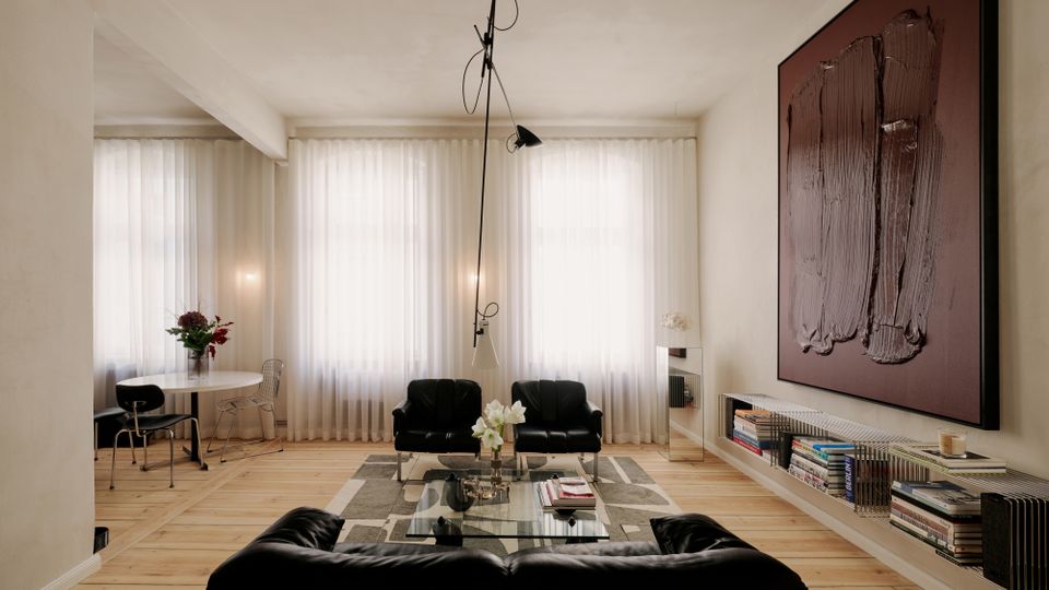 Helles, modernes Wohnzimmer mit breiten Dielenböden, großen Fenstern mit weißen Vorhängen und einer schwarzen Sitzgruppe um einen gläsernen Couchtisch auf einem gemusterten Teppich. Rechts hängt ein großes abstraktes Wandbild über einem niedrigen Regal mit Büchern, links steht ein kleiner runder Esstisch mit Stühlen.