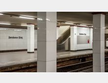 U-Bahn-Station am Potsdamer Platz in Berlin mit weiß gefliesten Wänden, schwarzen Schriftzügen mit Stationsnamen, Säulen sowie einem Rolltreppenaufgang. Im Vordergrund sind die Gleise und die Bahnsteigkante sichtbar.