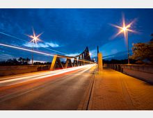 Eine beleuchtete Brücke bei Dämmerung, mit einer modernen Stahlkonstruktion und geschwungenem Design. Im Vordergrund sind Lichtspuren vorbeifahrender Fahrzeuge sichtbar, sowie Gehwege mit Backsteinpflaster und Straßenlaternen entlang der Brücke.