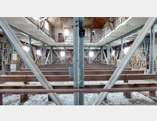 Innenansicht eines Dachbodens mit freigelegtem Fachwerk aus Holz- und Stahlträgern. Der Raum zeigt unverputzte Ziegelwände, Stahlstützen zur Stabilisierung und eine Dachkonstruktion mit Holzverkleidung, während unter den Trägern Isoliermaterial sichtbar ist.