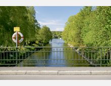Blick von einer Brücke auf einen Fluss, der von Bäumen gesäumt ist. Im Vordergrund ein grünes Metallgeländer mit einem Rettungsring und Hinweisschild befestigt.