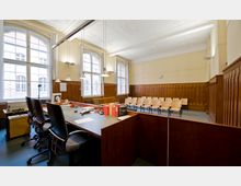 Ein Sitzungsraum mit hohen Decken, großen Fenstern und Holzwandvertäfelung. Im Vordergrund stehen Schreibtische mit Bürostühlen und Büromaterial, im Hintergrund befinden sich Holzstühle für Publikum oder Teilnehmer.