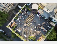 Luftaufnahme einer begrünten Dachterrasse mit einer Veranstaltung. Zu sehen sind Sitzreihen, Stehtische, mehrere Personen, ein Buffet, Sonnenschirme sowie umliegende Gebäude und eine Straße mit geparkten Autos.