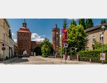 Straßenszene mit einem historischen Stadttor aus roten Backsteinen und einem Rundturm, flankiert von Wohn- und Geschäftshäusern. Im Vordergrund steht ein großes Banner für ein Fest, während links Autos und Fußgänger zu sehen sind.