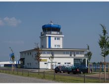 Flughafengebäude mit einem weißen Kontrollturm, der oben eine blaue Glaskuppel besitzt. Vor dem Gebäude sind geparkte Autos, junge Bäume entlang eines Gehwegs und ein eingezäuntes Rollfeld im Hintergrund sichtbar.