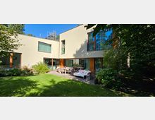 Modernes Wohnhaus mit zwei Etagen und flacher Fassade, großen Fenstern mit Holzrahmen und einer Terrasse, die mit Ess- und Sitzmöbeln ausgestattet ist. Im Vordergrund ein gepflegter Rasen und umliegende Bepflanzung, im Hintergrund Bäume.