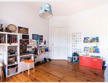 Helles Kinderzimmer mit Holzboden, weißen Wänden und Spielzeugregalen. An der Wand befinden sich bunte Buchstaben und Zahlen, Regale mit Kinderbüchern sowie Bilder und Poster; eine rote Spielzeugtruhe und ein hellblauer Lampenschirm mit Wolkenmotiv ergänzen die Einrichtung.