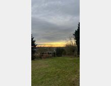 Weite, grüne Wiese mit vereinzelten Bäumen und Sträuchern, im Hintergrund ein kleines, von Bäumen umgebenes Gartenhäuschen. Am Horizont sichtbar ist ein gelb-orange leuchtender Abendhimmel unter einer dichteren Wolkendecke.