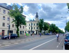 Straßenansicht mit einer breiten, von Bäumen gesäumten Straße, auf der Radfahrer unterwegs sind. Im Hintergrund befindet sich ein historisches Gebäude mit einer verzierten weißen Fassade und einem Turm mit Kuppelspitze.