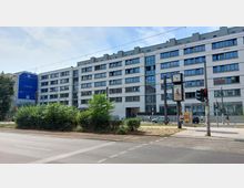 Moderne mehrstöckige Gebäudezeile mit weißer Fassade und zahlreichen Fenstern, im Vordergrund eine Grünfläche, eine Straßenkreuzung und eine Uhr mit Werbetafel. Fußgänger und Radfahrer sind entlang des Gebäudes sichtbar, rechts befindet sich eine Ampel.