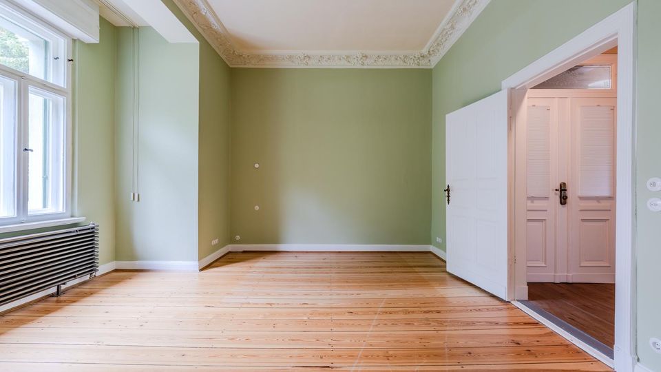 Leerer Wohnraum mit hellgrünen Wänden, hohen Fenstern und einem Heizkörper unter dem Fenster sowie breiten Holzdielenboden. Stuckverzierte Deckenleisten und eine weiße Tür führen in einen angrenzenden Flur mit weiteren Türen.