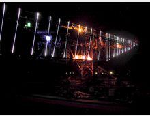 Industrielle Stahlkonstruktion einer Brücke oder eines Krans, nachts beleuchtet durch bunte LED-Lichter in Blau, Gelb und Weiß, die dünne, vertikale Linien bilden. Die Umgebung ist dunkel, und die Konstruktion hebt sich durch die helle Beleuchtung deutlich ab.