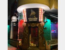 Eingang zu einer Bar oder einem Club mit dem Namen „Süß war gestern“, umgeben von farbigen Neonlichtern in Rot und Grün. Der Eingang ist dekoriert mit einem hölzernen Portal mit geschwungenen Mustern und einer illuminierten Beschriftung sowie einem Schild mit einem bewaffneten Kaninchen.