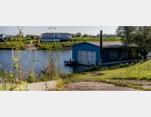 Ein Uferbereich mit einem blauen Holzgebäude direkt am Wasser, das ein Garagentor und Sprossenfenster hat. Im Hintergrund befindet sich ein größeres weiß-blaues Gebäude mit Fahnenmasten, das auf einem begrünten Deich steht.