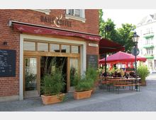 Ein Café in einem Backsteingebäude mit großen Holzfenstern und einer roten Markise, umgeben von Pflanzkübeln mit Bambus. Vor dem Café stehen im Außenbereich Tische und Sitzbänke unter roten Sonnenschirmen, mit weiteren Wohngebäuden im Hintergrund.