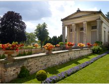 Klassizistisches Gebäude mit Säulenfassade, umgeben von einer gepflegten Gartenanlage mit blühenden Blumen in Steinschalen auf einer niedrigen Backsteinmauer. Im Hintergrund sind Bäume und ein leicht bewölkter Himmel zu sehen.