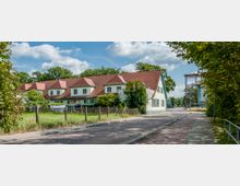 Straßenszene mit einem Wohngebäude im Landhausstil, das grüne Wände, rote Ziegeldächer und weiße Fensterläden hat. Im Vordergrund sind eine gepflegte Wiese, ein Zaun und ein Gehweg zu sehen; rechts befindet sich eine Bauschleuse oder ein Turmgerüst, umgeben von Bäumen und Grünflächen.