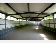 Überdachter Fußgängertunnel mit Metallkonstruktion und durchscheinenden Wänden, ausgestattet mit Geländern auf beiden Seiten und einem Betonboden. Der Tunnel führt geradeaus, während Tageslicht durch die Seitenfenster fällt.