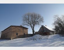 Verschneiter Hof mit zwei ländlichen Gebäuden, links ein Backsteinbau und rechts ein älteres Haus mit Satteldach. Dazwischen steht ein großer, kahler Baum auf einer schneebedeckten Böschung unter klarem Himmel.