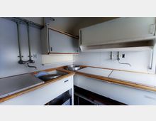 Kleine, funktionale Küchenzeile in einem einfachen Raum mit weißer Ausstattung, Holzdetails und Metallarmaturen. Sichtbar sind mehrere Spülbecken, Schränke und Rohre an der Wand.